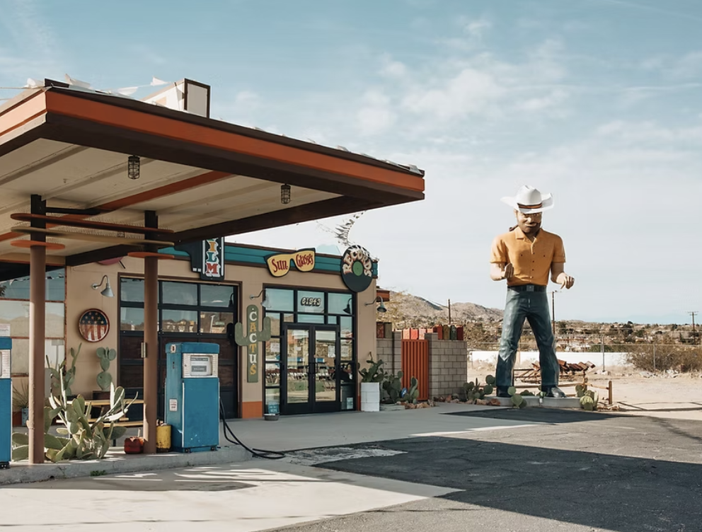 A classic American roadside diner with cactus plants outside, featuring a large cowboy statue wearing a white cowboy hat, orange shirt, and jeans, with a desert landscape in the background.