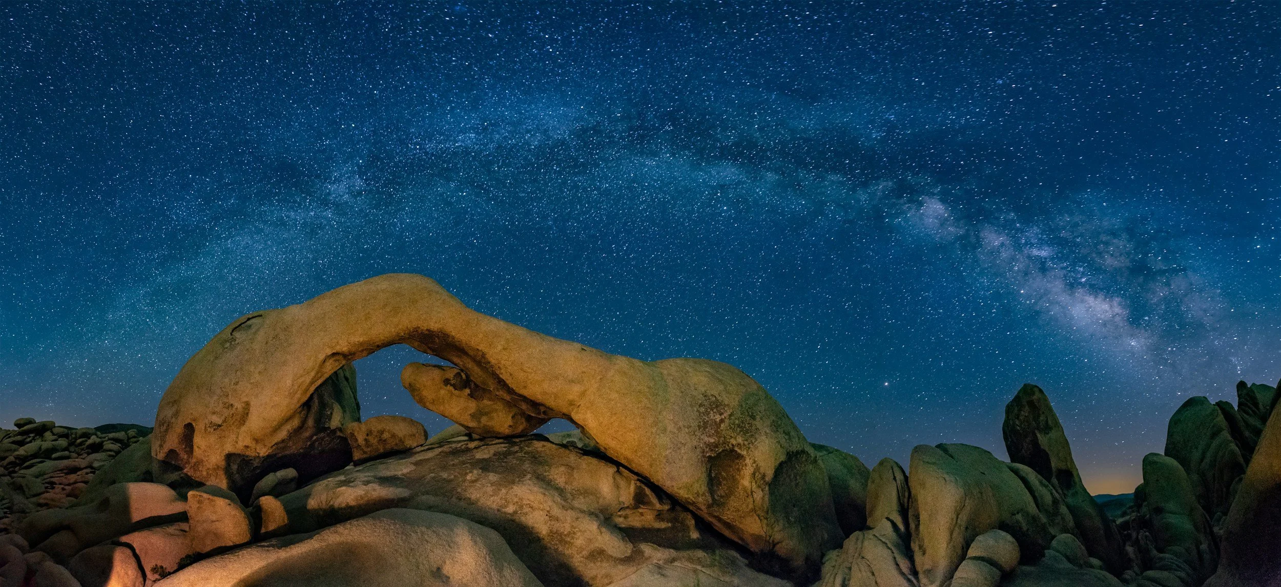 Starry night sky with the Milky Way galaxy above large, uniquely shaped rocks in a desert landscape.