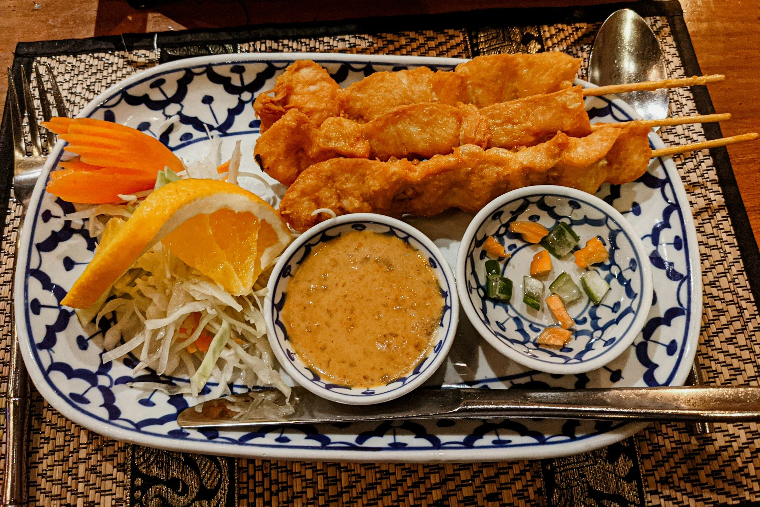 Plate of Thai food with grilled chicken skewers, shredded cabbage salad with orange slices, and two dipping sauces on a patterned ceramic plate.