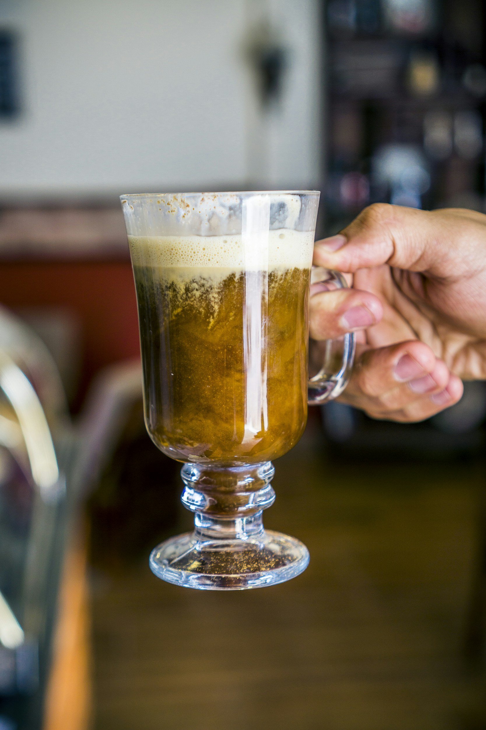 A hand holding a glass mug filled with beer, showing foam on top and bubbles inside, in a cozy indoor setting.