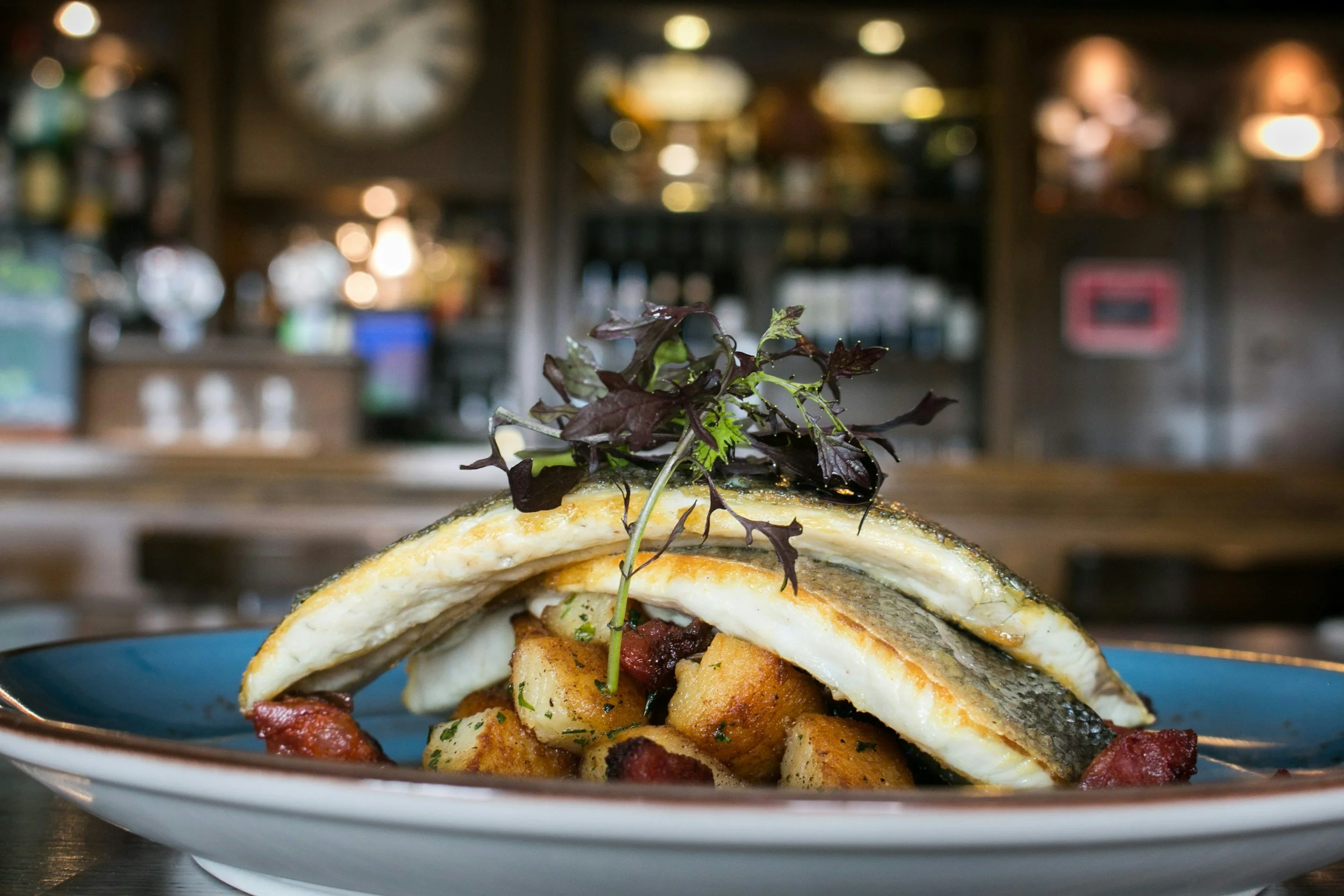 Fried fish fillets served on a plate with roasted potatoes, garnished with microgreens, in a cozy restaurant setting.