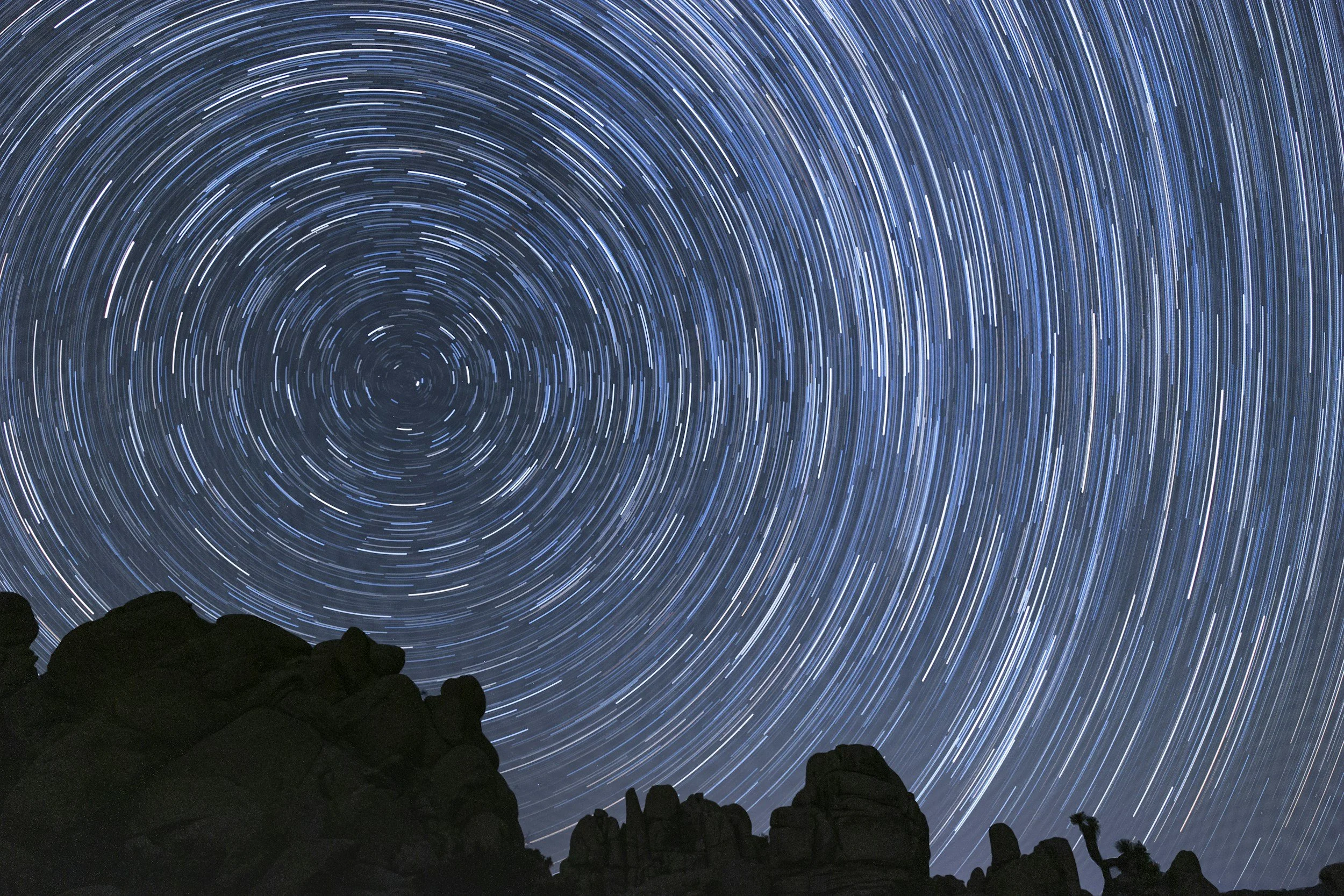 Long exposure photograph of star trails in the night sky above a silhouette of rocky landscape.