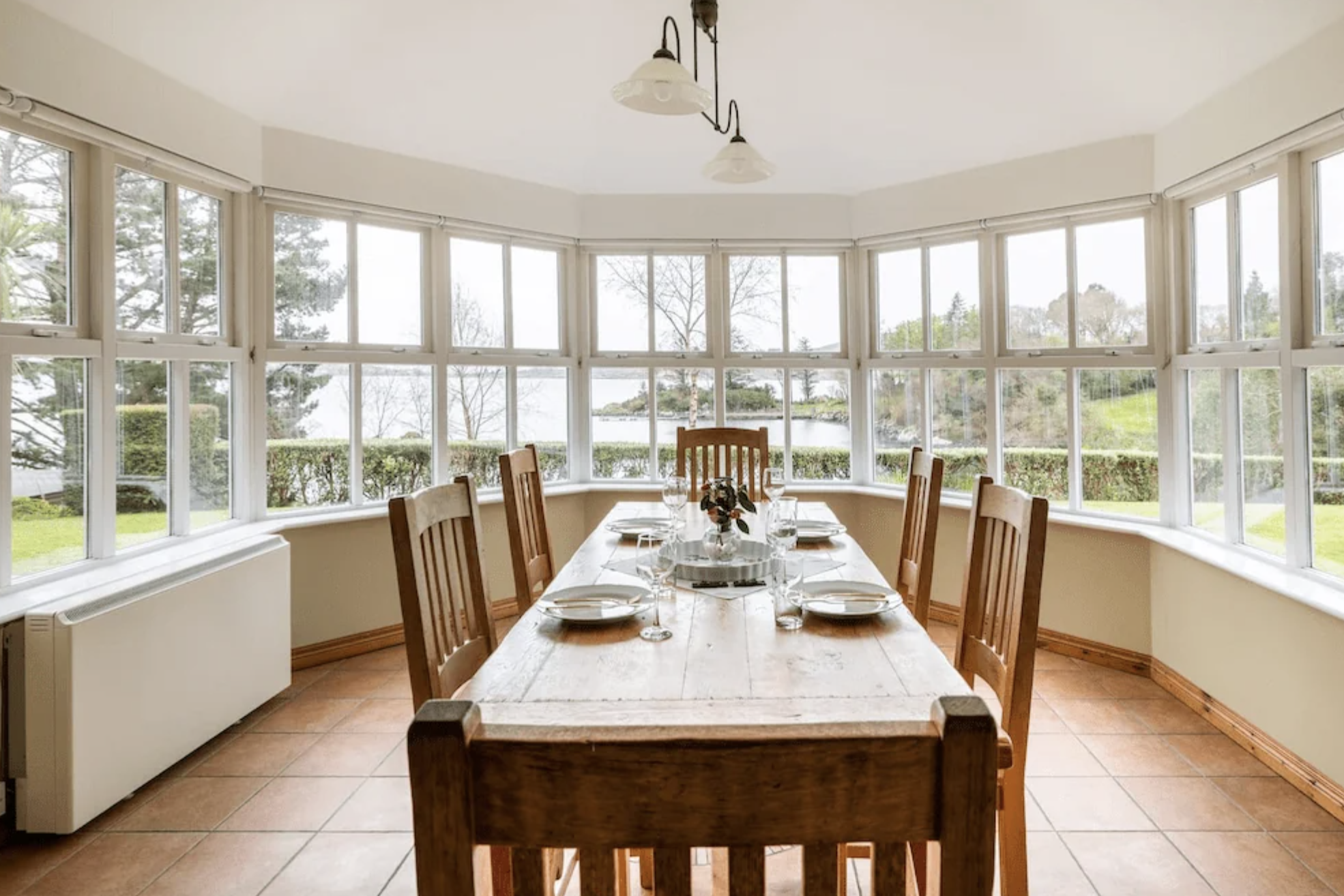 Dining room with large windows showing a view of water and greenery, wooden dining table set with plates, glasses, and a vase with flowers, and a chandelier overhead at the location of a yoga and art retreat in Ireland.