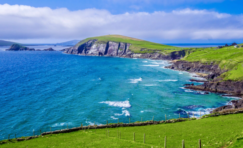 Scenic coastal landscape with green grassy hills, rocky cliffs, and deep blue ocean under partly cloudy sky.
