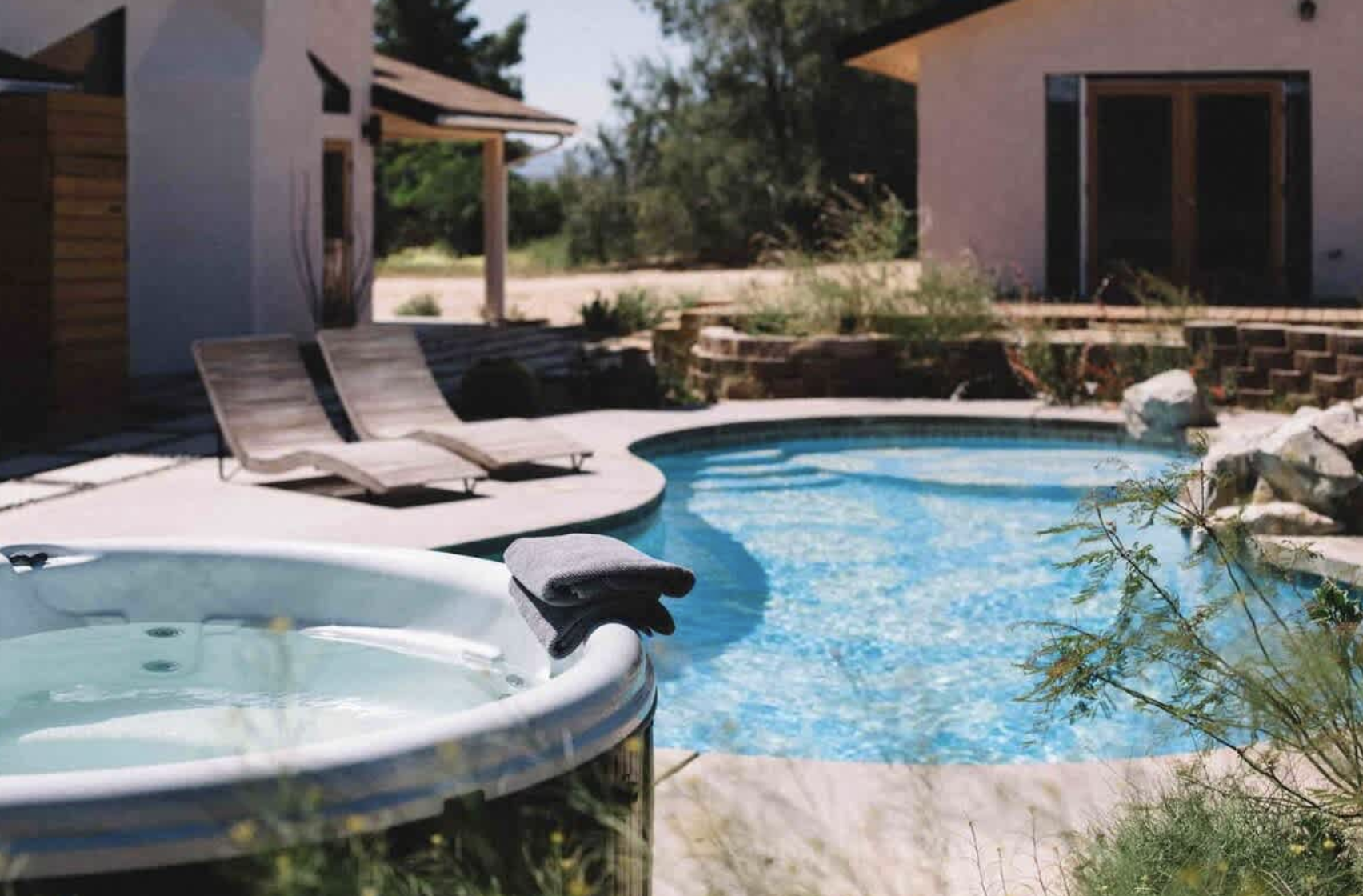 An outdoor swimming pool area with lounge chairs, a hot tub, and a modern house in the background, surrounded by trees and patio landscaping.