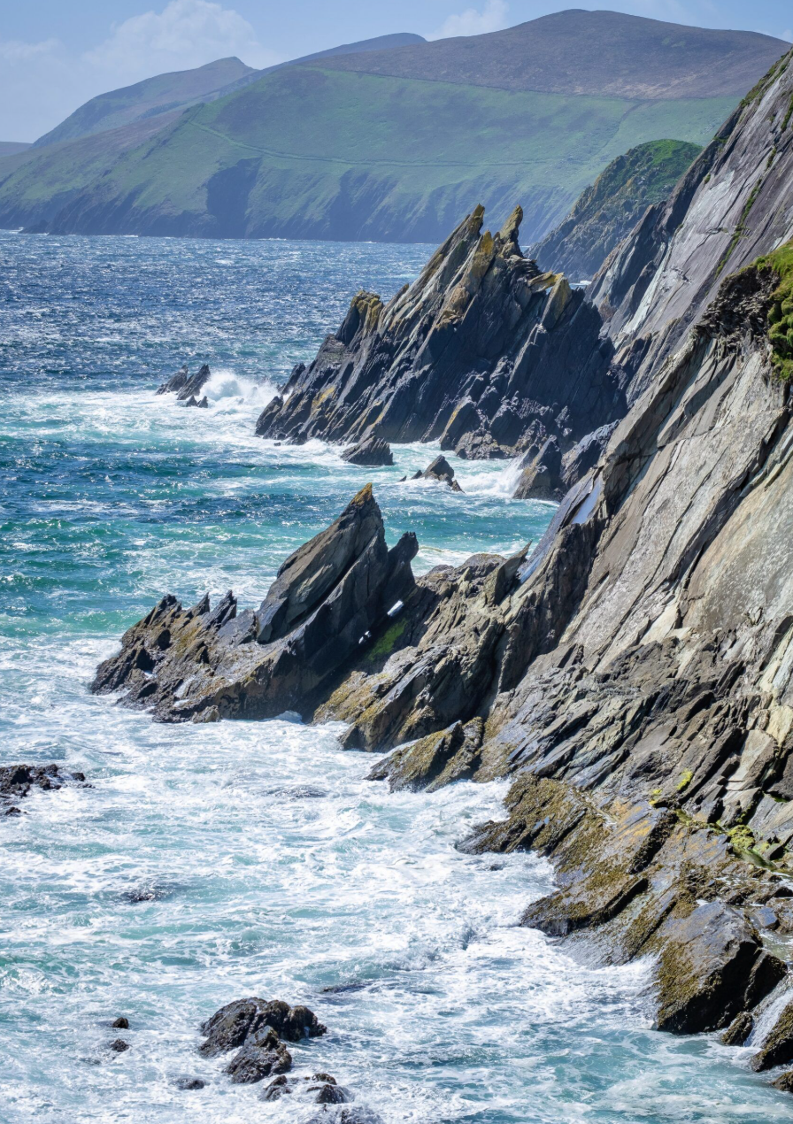 A rugged coastline with jagged rock formations extending into the ocean, with waves crashing against them, and lush green hills in the background under a cloudy sky.