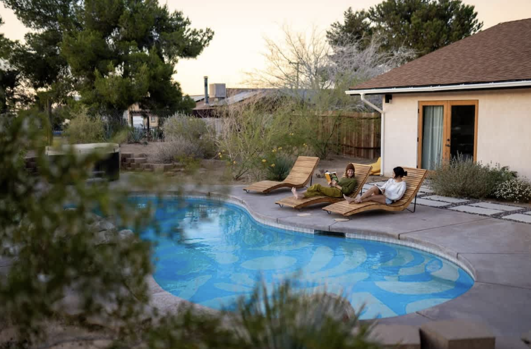 Two women relaxing on wooden lounge chairs by a swimming pool at sunset in a backyard, with trees and a house in the background.
