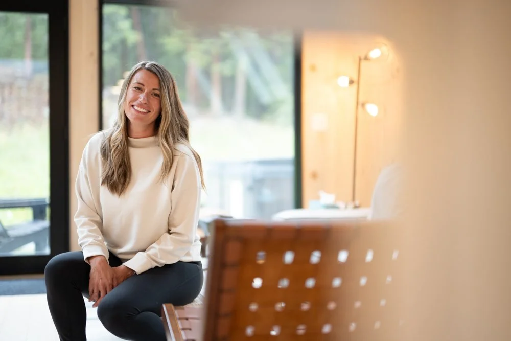 A woman with long wavy hair, wearing a white sweatshirt and black pants, sitting on a bench in a well-lit indoor space with large windows and a view of greenery outside.