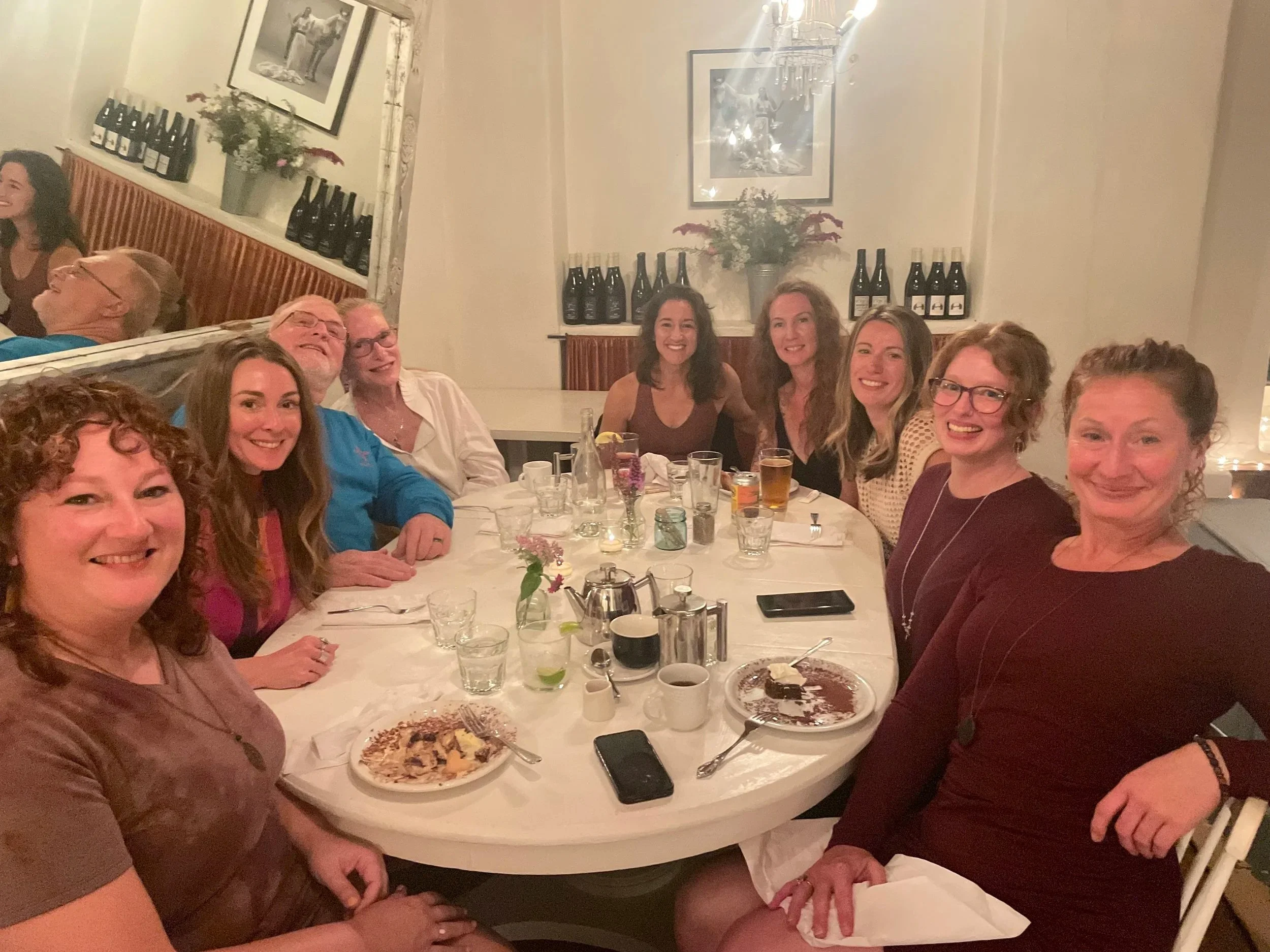 A group of nine women sitting around a table at a dinner party, smiling for a photo in a decorated indoor setting with bottles on the wall and floral arrangements.