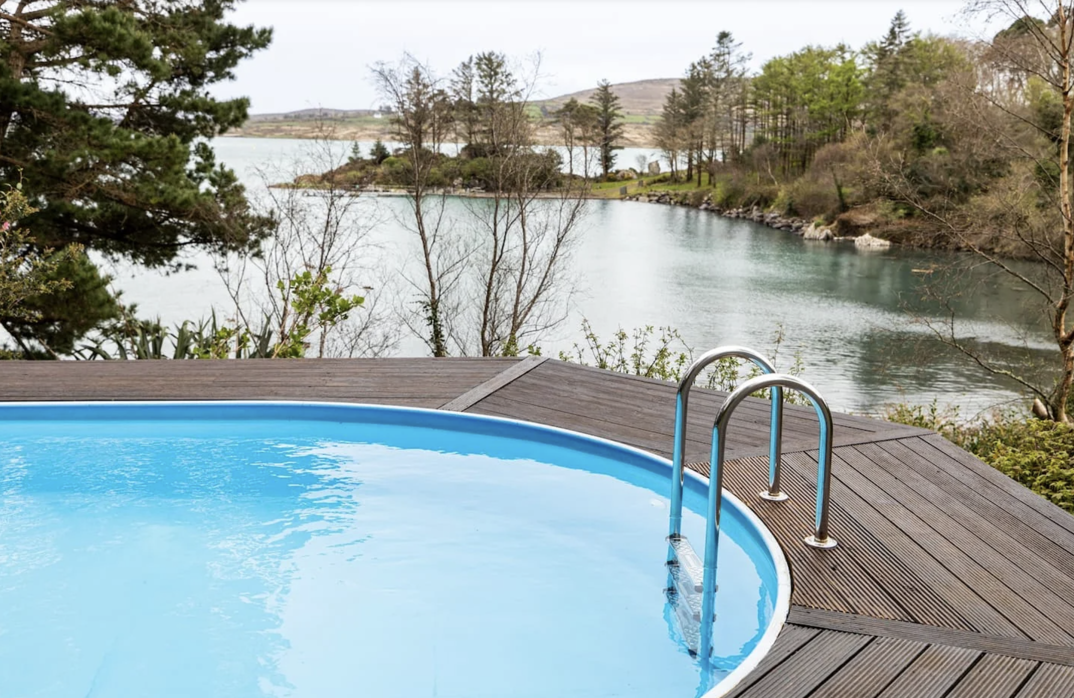 An outdoor hot tub with blue water on a wooden deck overlooking a river with trees and hills in the distance at the location of a yoga and art retreat in Ireland.