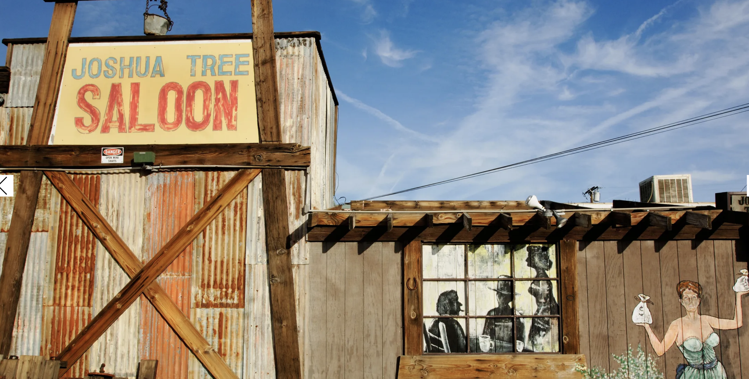 A rustic building with a sign that reads "Joshua Tree Saloon" in blue and red lettering, and a mural of three women with drinks, a horsehoe, and a woman holding a bag of money outside.