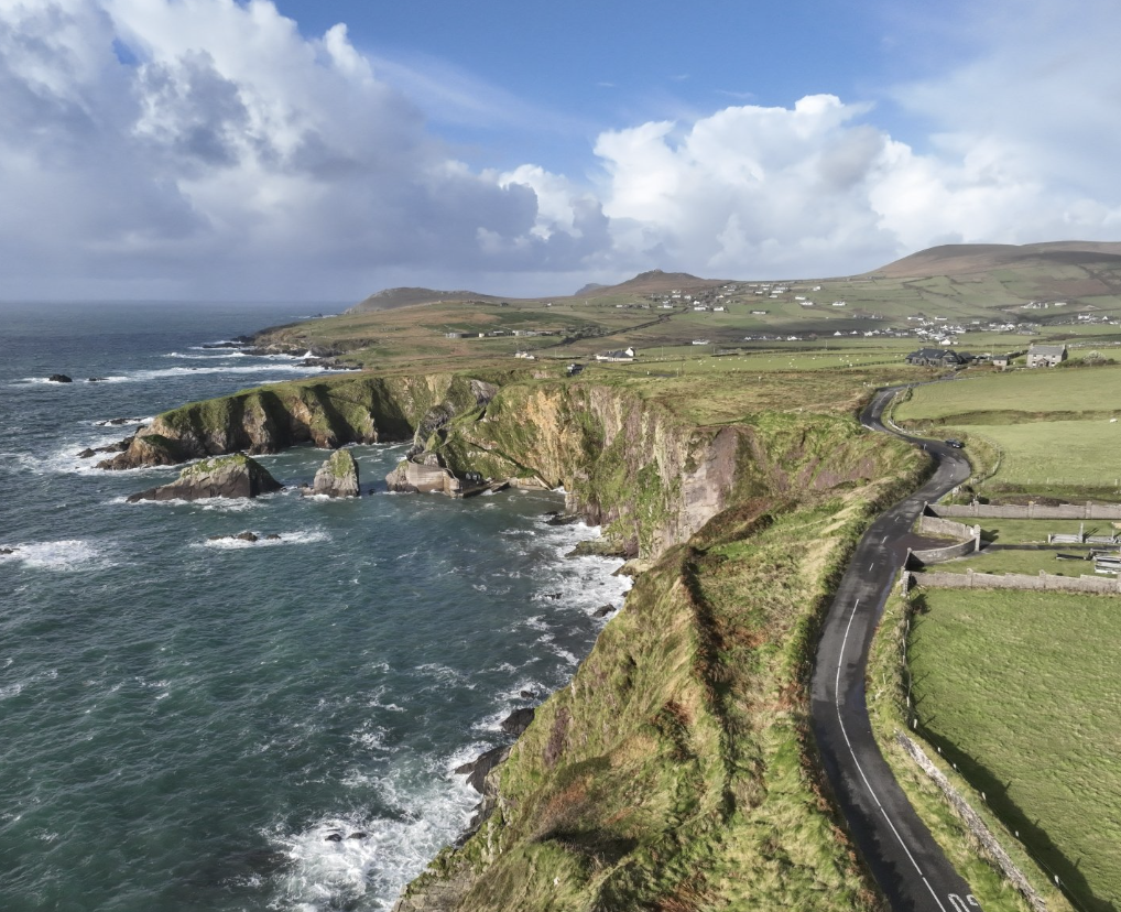 A coastal landscape with steep green cliffs, a winding road, green fields, and white houses overlooking the ocean under a partly cloudy sky.