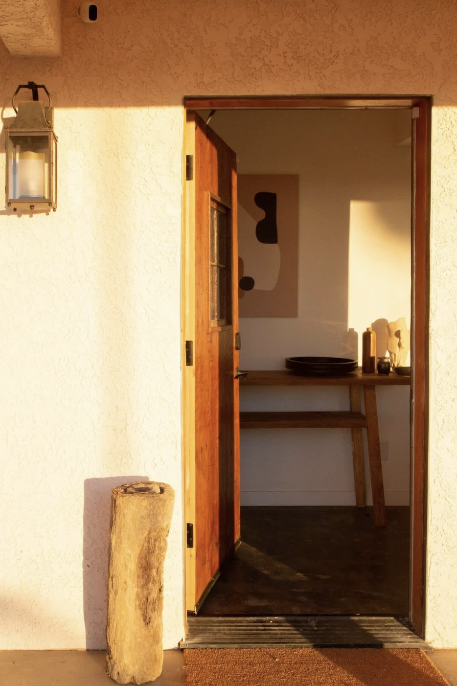 The image shows the open door of a house entrance with warm sunlight casting light on the wall and door. There is a rustic wooden log placed on the ground outside, and inside, a wooden table with decorative items, including plates and vases, is visible.
