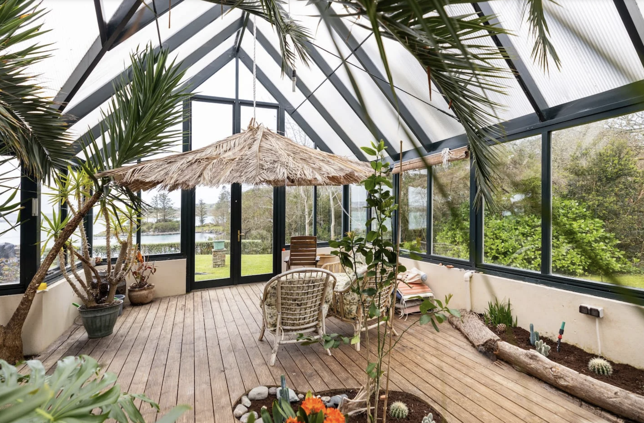 A glass-enclosed sunroom with wooden flooring, decorated with potted plants, a thatched umbrella, and outdoor furniture, overlooking a lush green landscape with water in the distance where yoga will take place.
