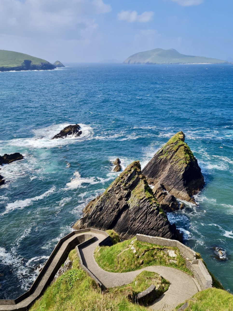 A scenic coastal view showing a rocky shoreline with large jagged rocks and ocean waves, a walkway winding along the cliffside with green grass, and distant islands or landforms under a partly cloudy sky.