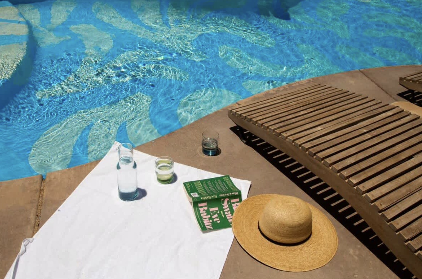 Poolside scene with a white towel, three glasses of water, a book, and a straw hat on the concrete surface next to a swimming pool with clear blue water.