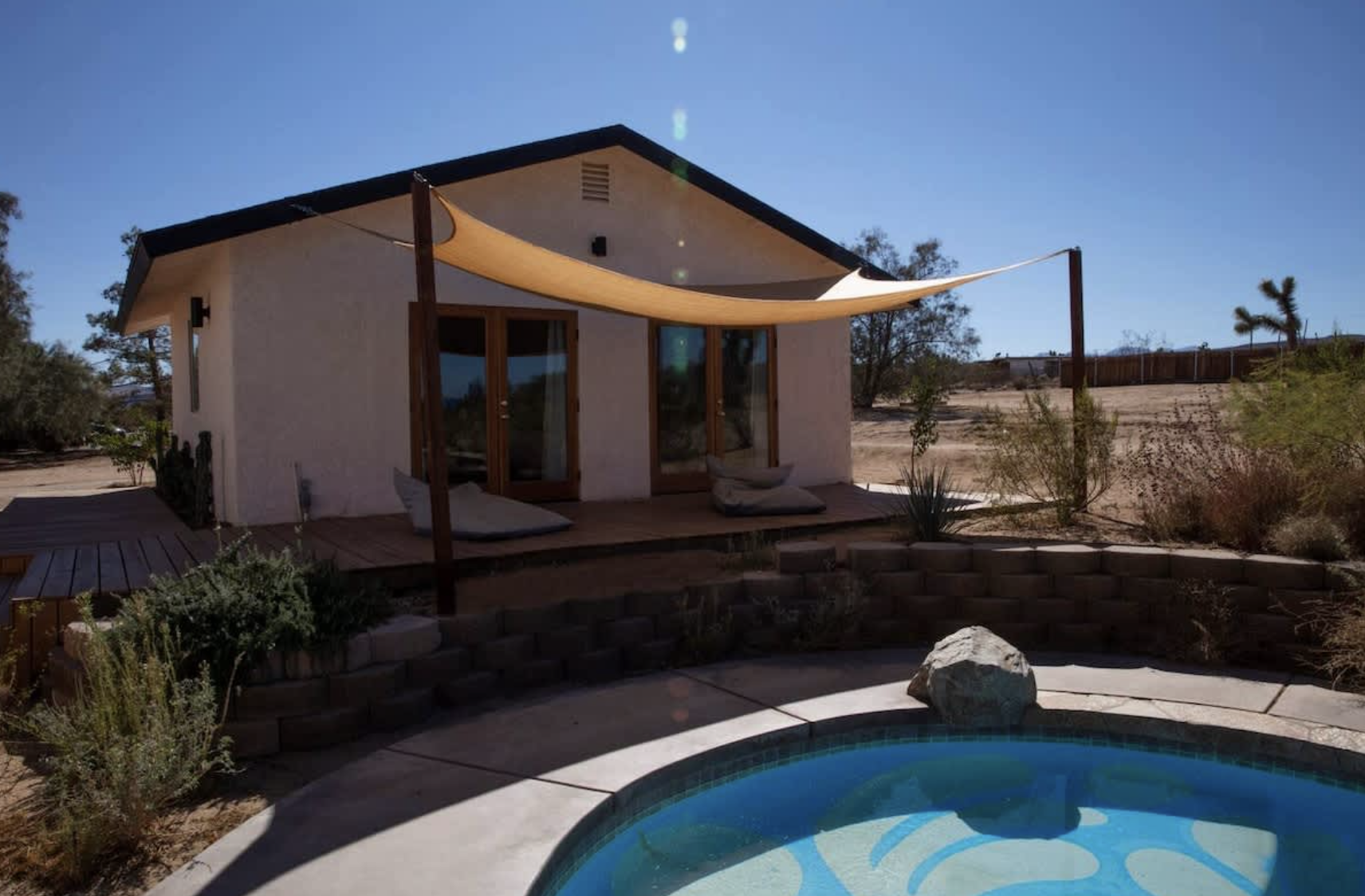 A small house with a wooden deck, two glass doors, and a shade sail on the patio. In front, there is a swimming pool with a large rock in the corner. Desert plants and trees are in the background under a clear blue sky.
