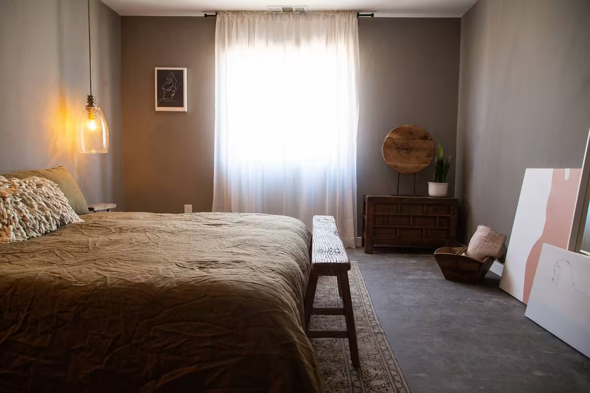 A cozy bedroom with a large bed, textured pillows, a hanging pendant light, a framed artwork, a window with sheer curtains, a wooden bench at the foot of the bed, a wooden dresser with a round mirror and potted plant, a basket with cushions, and two abstract art pieces leaning against the wall.