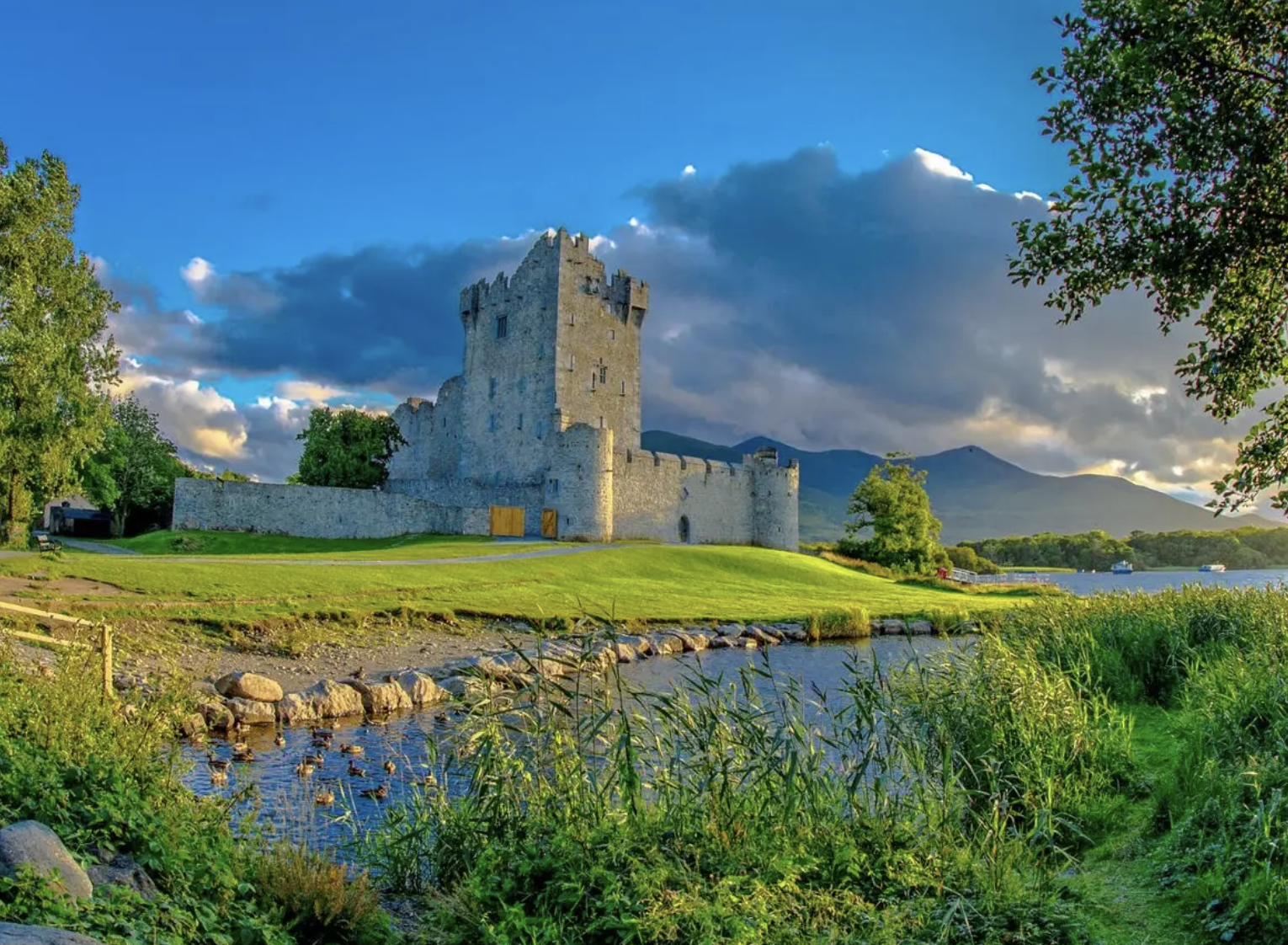 A medieval Ireland castle beside a body of water with lush green grass and trees, mountains in the background, under a partly cloudy sky.