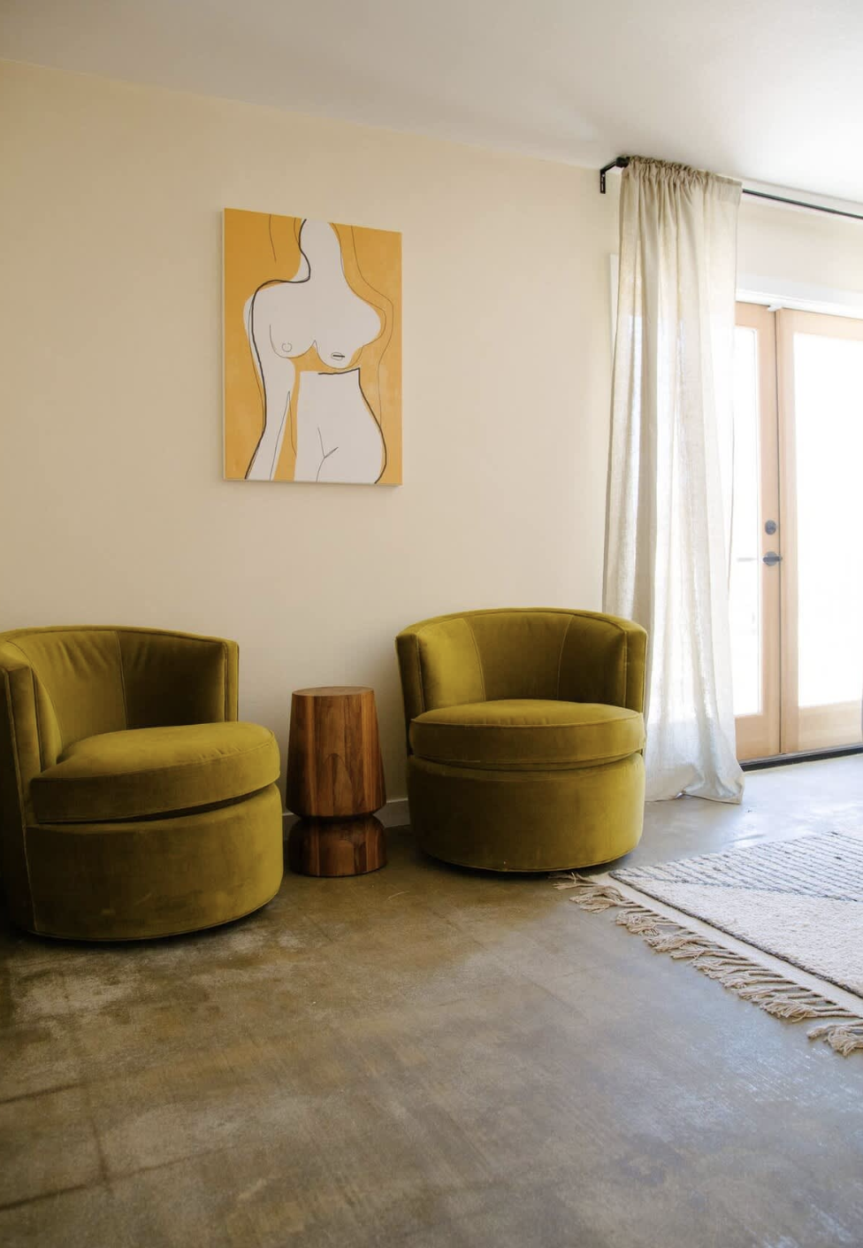 Living room with two green velvet armchairs, a small wooden side table between them, a framed line art painting of a female figure on the wall, cream curtains, and a glass door letting in natural light.