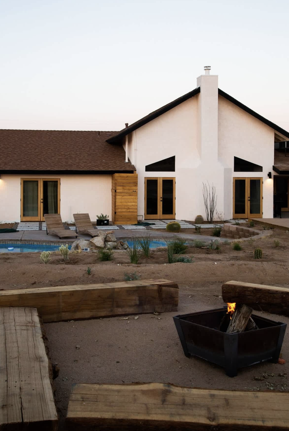 A modern house with large glass doors, a small swimming pool, lounge chairs, desert landscaping with cacti, and a fire pit in a backyard during sunset.