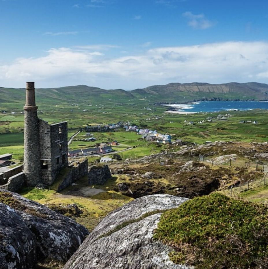 A scenic landscape in south Ireland with ruins of an old stone tower on a hill, green fields, a small coastal town, and mountains in the background under a partly cloudy sky.