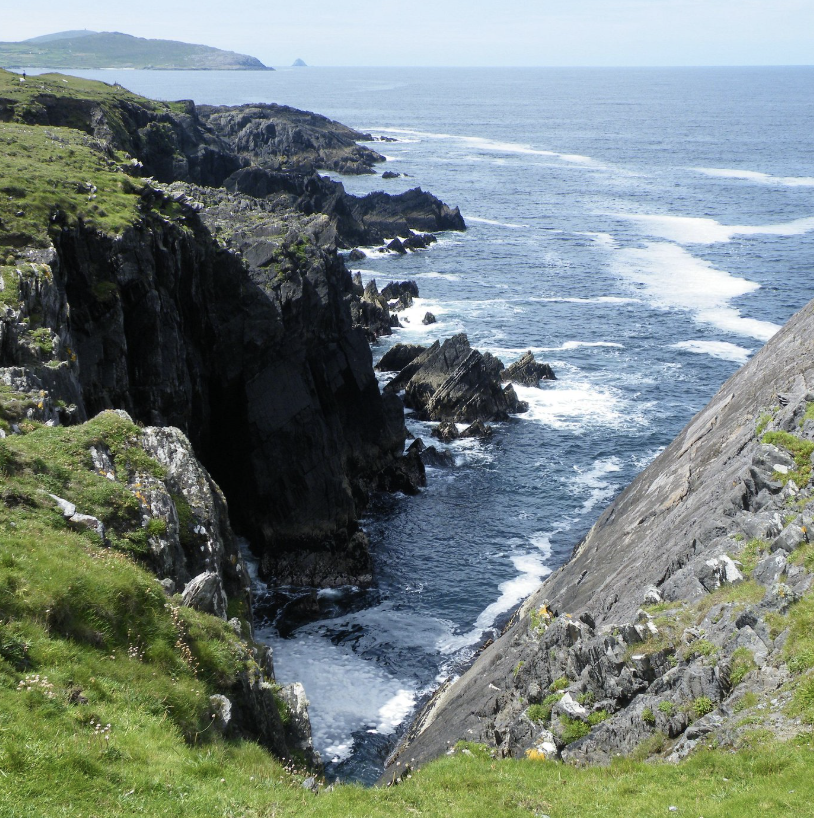 South Ireland cliffs along a rugged coastline with waves crashing at the base, green grass on top, and a distant landmass across the ocean.