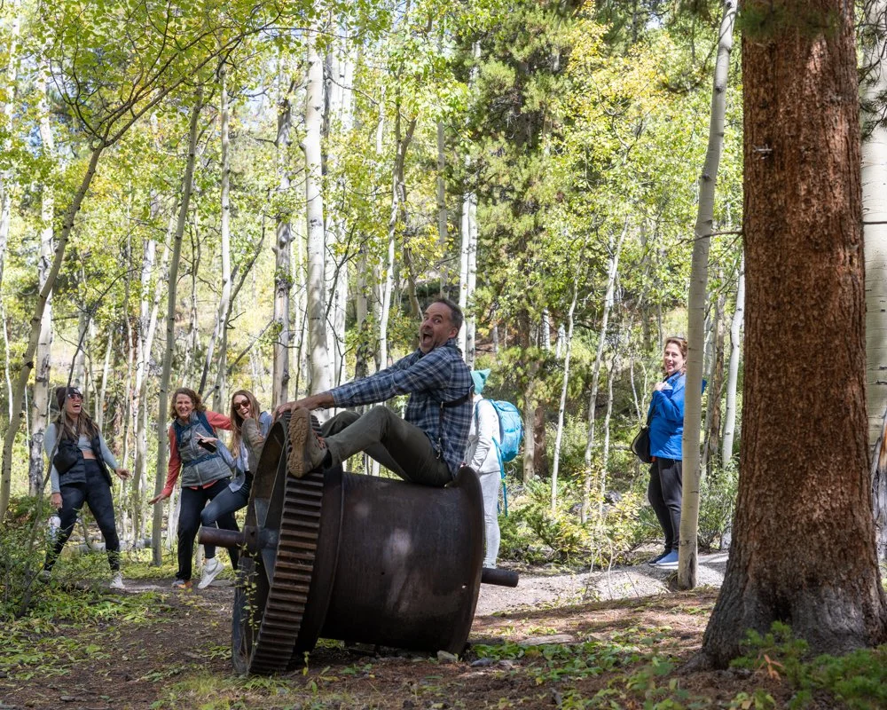A group of smiling people having fun outdoors in a forest, with a man sitting on an overturned large metal gear or machine part in the foreground.