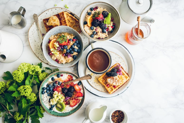 Breakfast table with bowls of oatmeal topped with berries and fruit, waffles, a cup of coffee, a glass jar of honey, and a potted plant on a white marble surface.