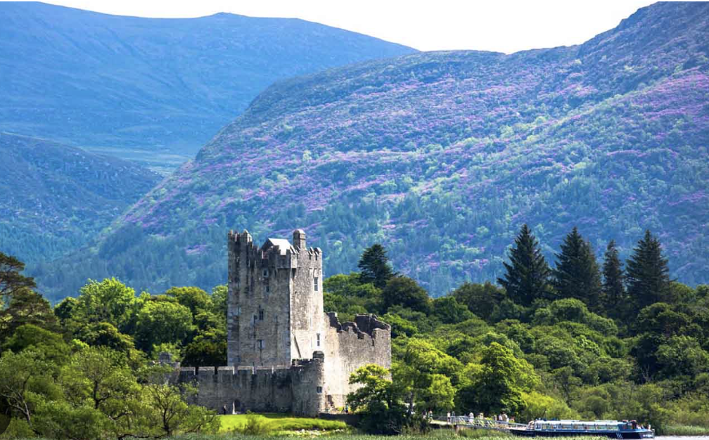 A castle surrounded by lush green trees with purple and green mountains in the background and a train passing by in the foreground in Killarney National Park in Ireland.