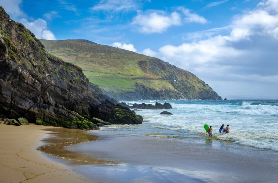 Beach with rocky cliffside, green hills, and people entering the ocean with surfboards