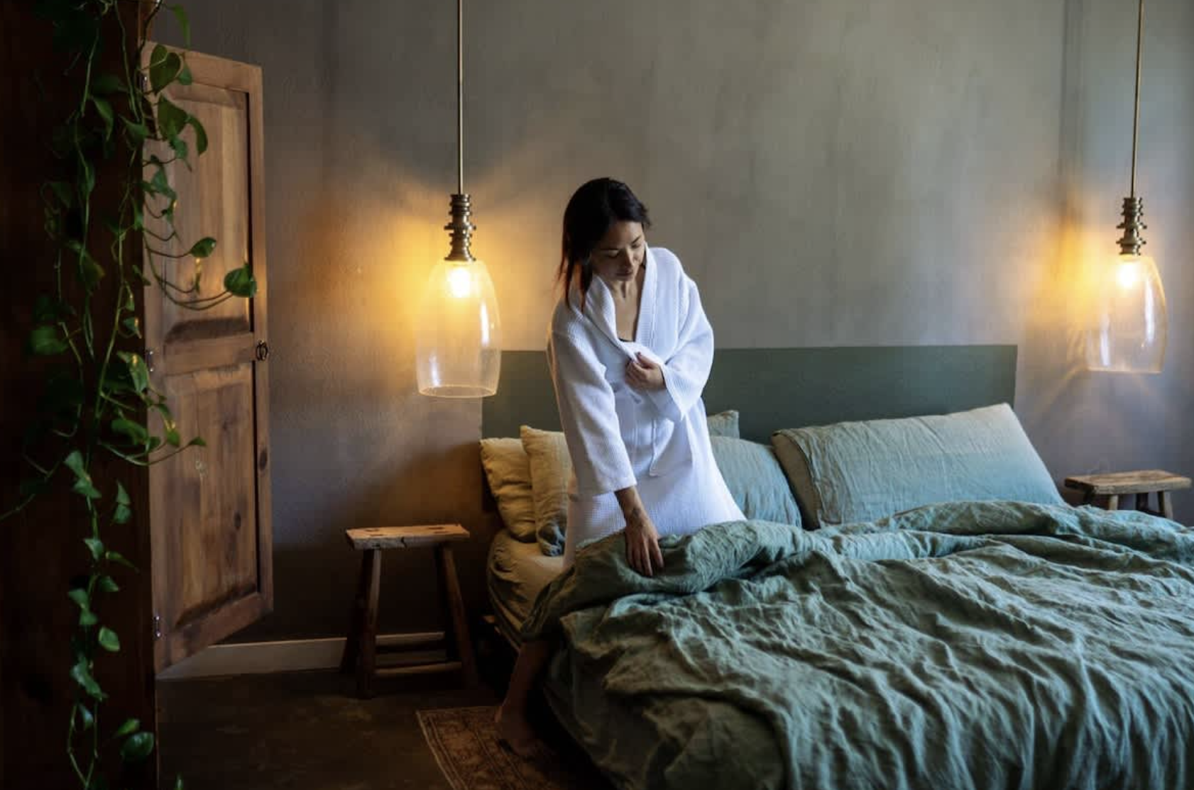 A woman in a white bathrobe making a bed in a dimly lit bedroom with hanging pendant lights and wooden furniture.