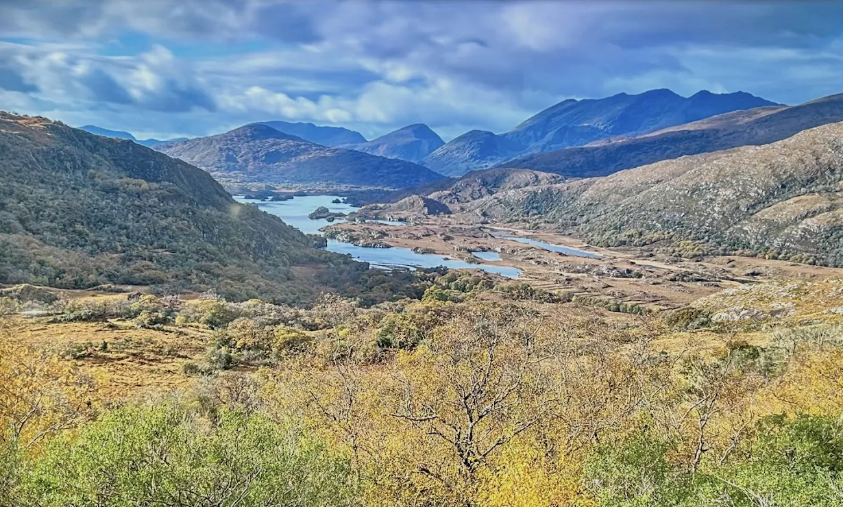 Scenic landscape of mountains, rivers, and rolling hills with trees in autumn colors under a partly cloudy sky in south Ireland.