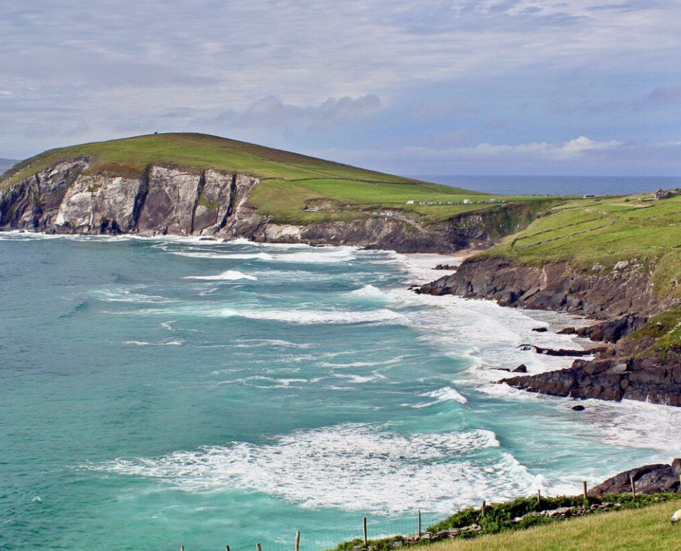 Scenic coastline with green hills, rocky cliffs, and turquoise ocean waves under a cloudy sky.