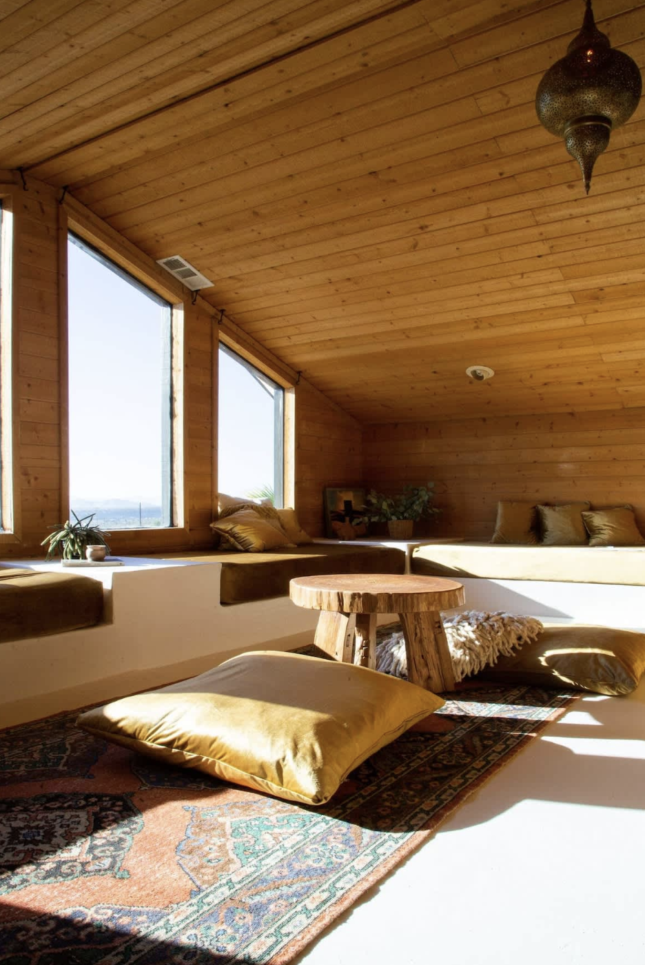 Sunlit cozy room with wooden walls and ceiling, large windows, cushions, and a small wooden table on a colorful patterned rug.