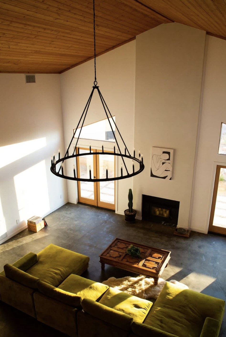 Living room with a yellow sofa, a wooden coffee table with a plant, a tall cactus, a fireplace, a modern black and white painting, large windows, and a high wooden ceiling with a circular chandelier.