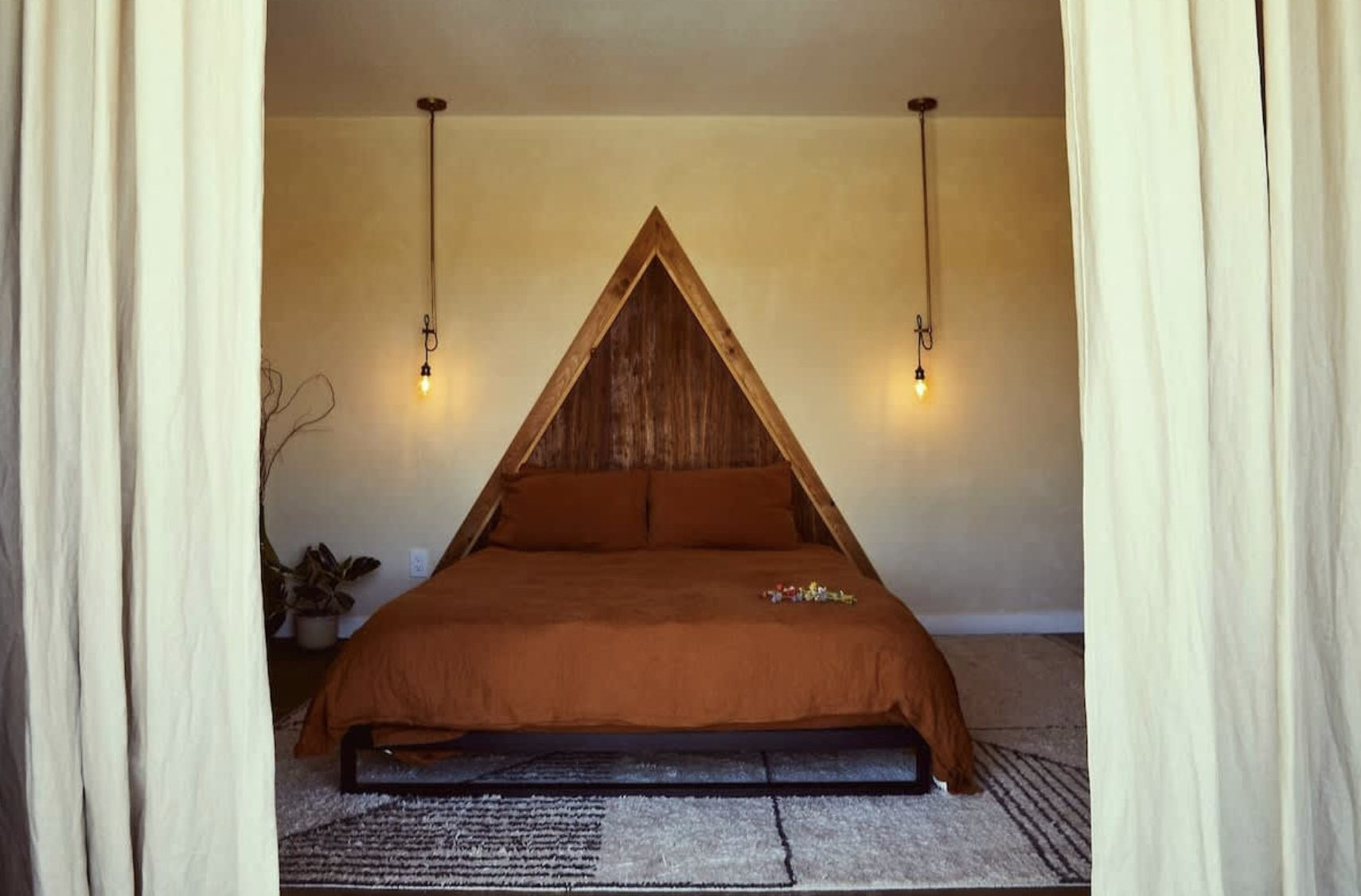 A bedroom with a bed that has a brown bedspread, positioned under a wooden triangular headboard. Two hanging pendant lights are on each side of the bed, and there is a small plant in a pot to the left.