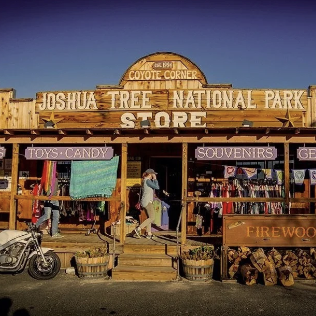 Front view of the Joshua Tree National Park Coyote Corner Store with wooden signage. The store sells toys, candy, souvenirs, and gear, with customers shopping outside. There is a motorcycle parked in front and wood logs stored to the side.