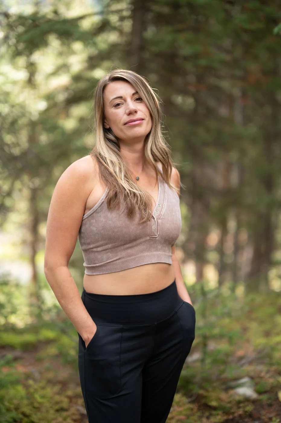 A woman standing outdoors in a forest, wearing a beige sleeveless crop top and black pants, with her hands in her pockets.