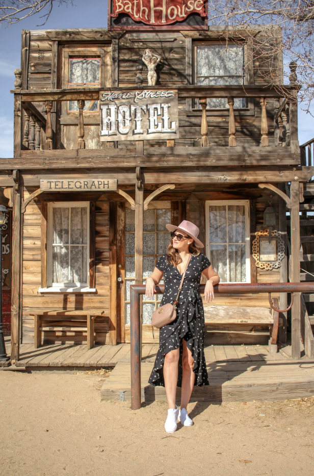 A woman in a black polka dot dress, pink hat, and sunglasses standing in front of a rustic wooden building with signs reading 'Home Street Hotel' and 'Telegraph,' in a Western town style.