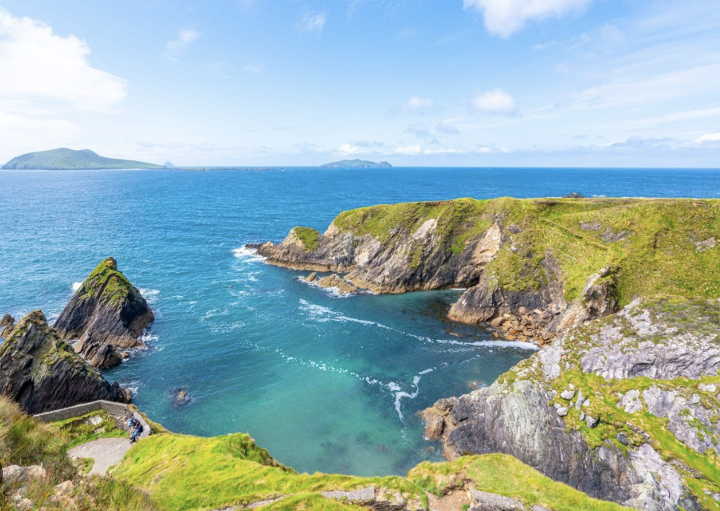A scenic view of a coastal landscape with rocky cliffs and green grass, overlooking the ocean with a blue sky and some clouds in the background.