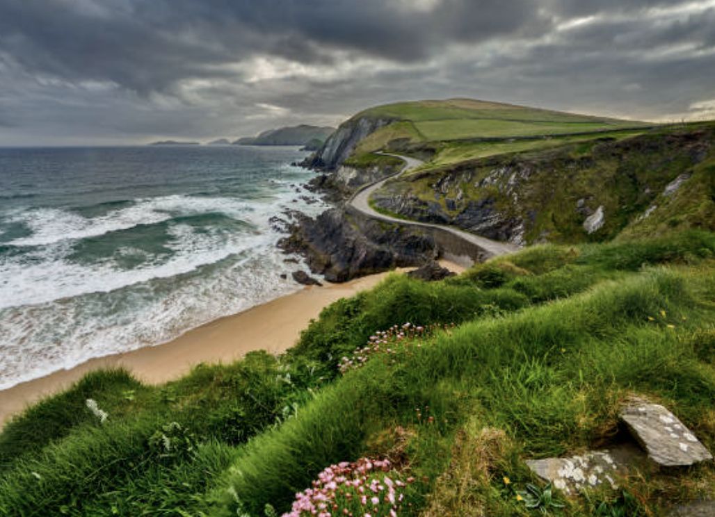 Coastal landscape with green cliffs, a winding road, and a sandy beach under a cloudy sky.