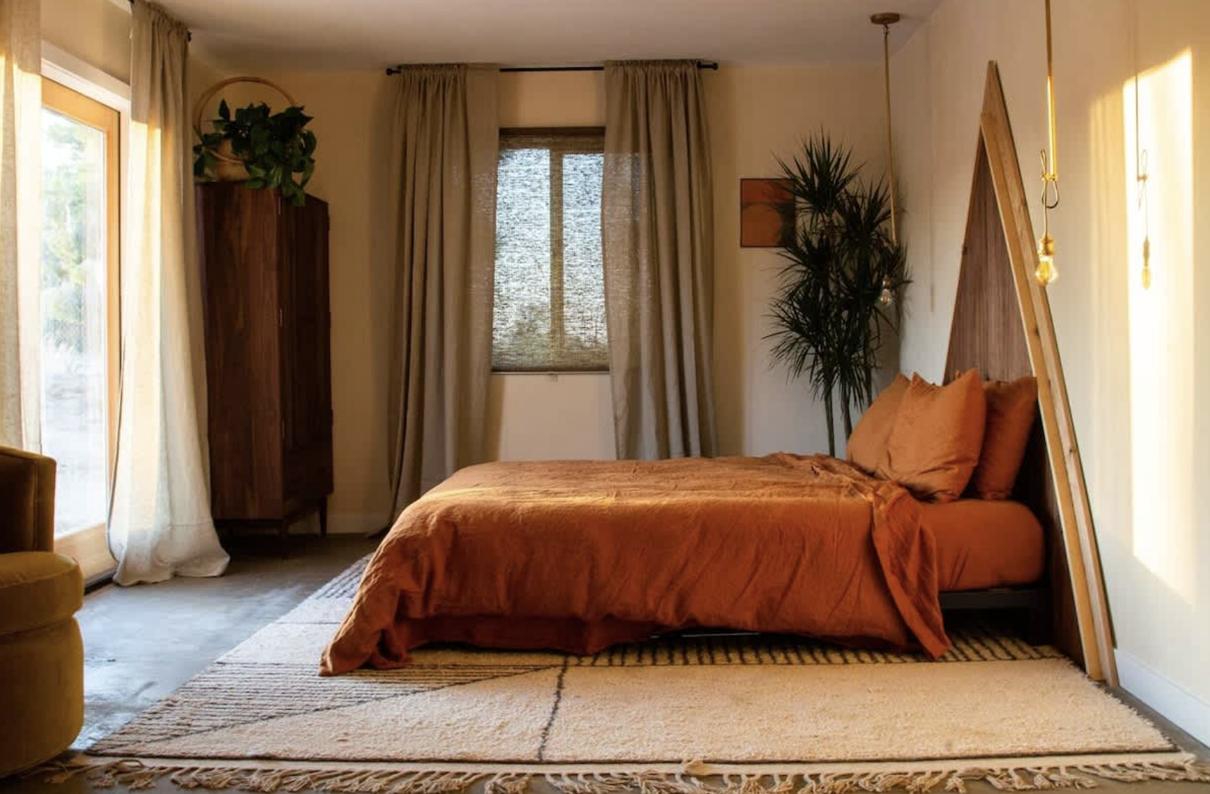 Interior of a bedroom with a bed covered in orange bedding, a wooden headboard with angled sides, a book on the bed, a tall wooden cabinet with a potted plant on top, a window with beige curtains, a sliding door with curtains, and hanging light bulbs.