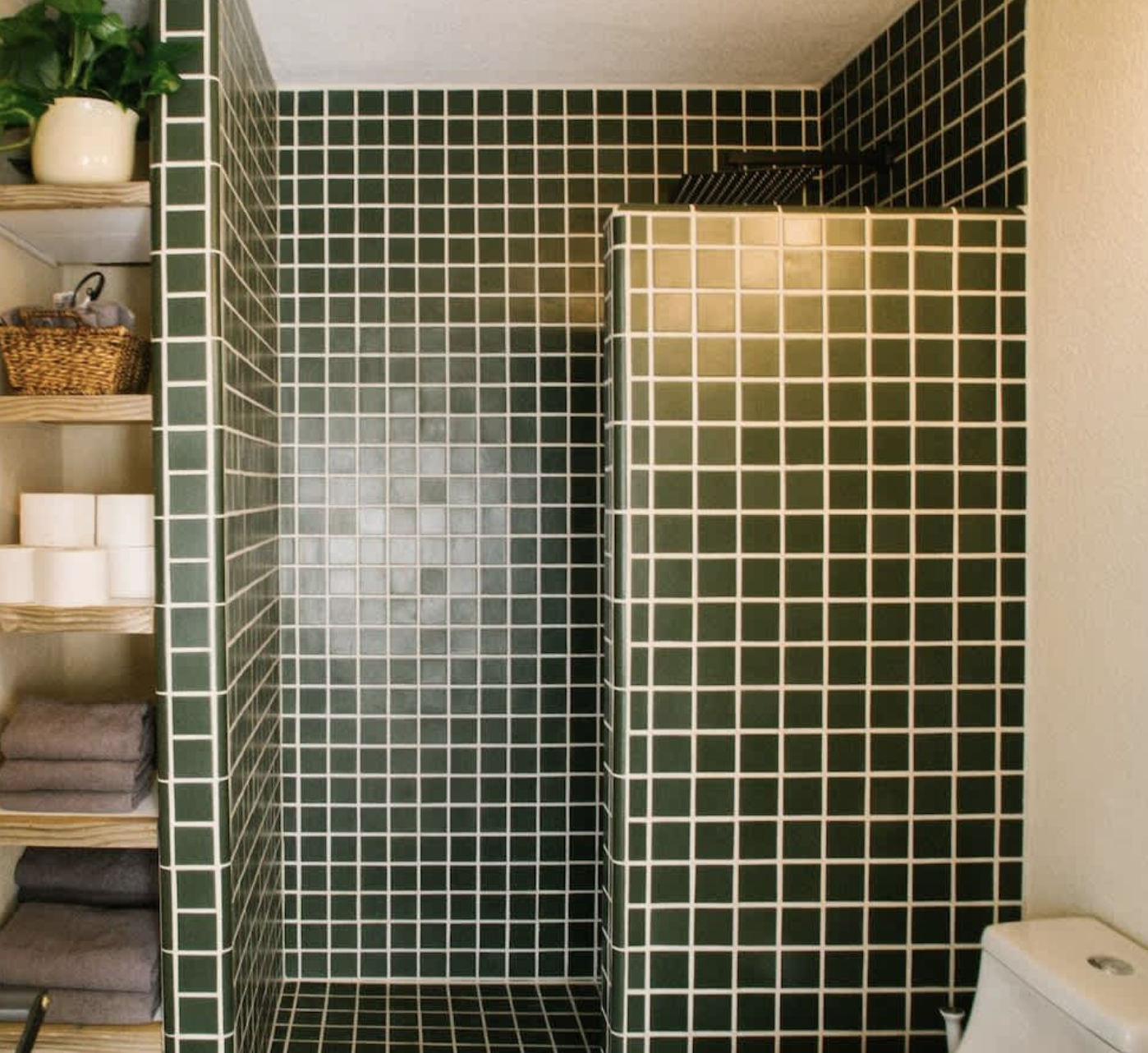 Green tiled shower with a partial view of a toilet on the right and a shelving unit with towels and toilet paper on the left side.