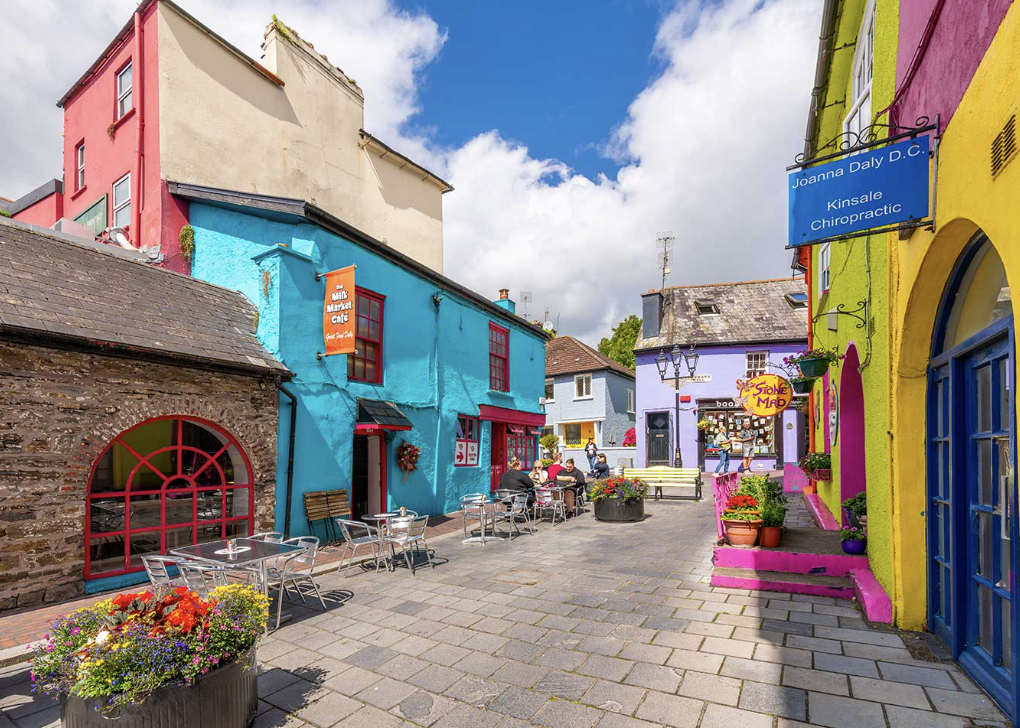 Colorful buildings lining a pedestrian street with outdoor seating, flowers, and a sign for a cafe at a bright blue building, under partly cloudy sky at a yoga retreat in south Ireland.