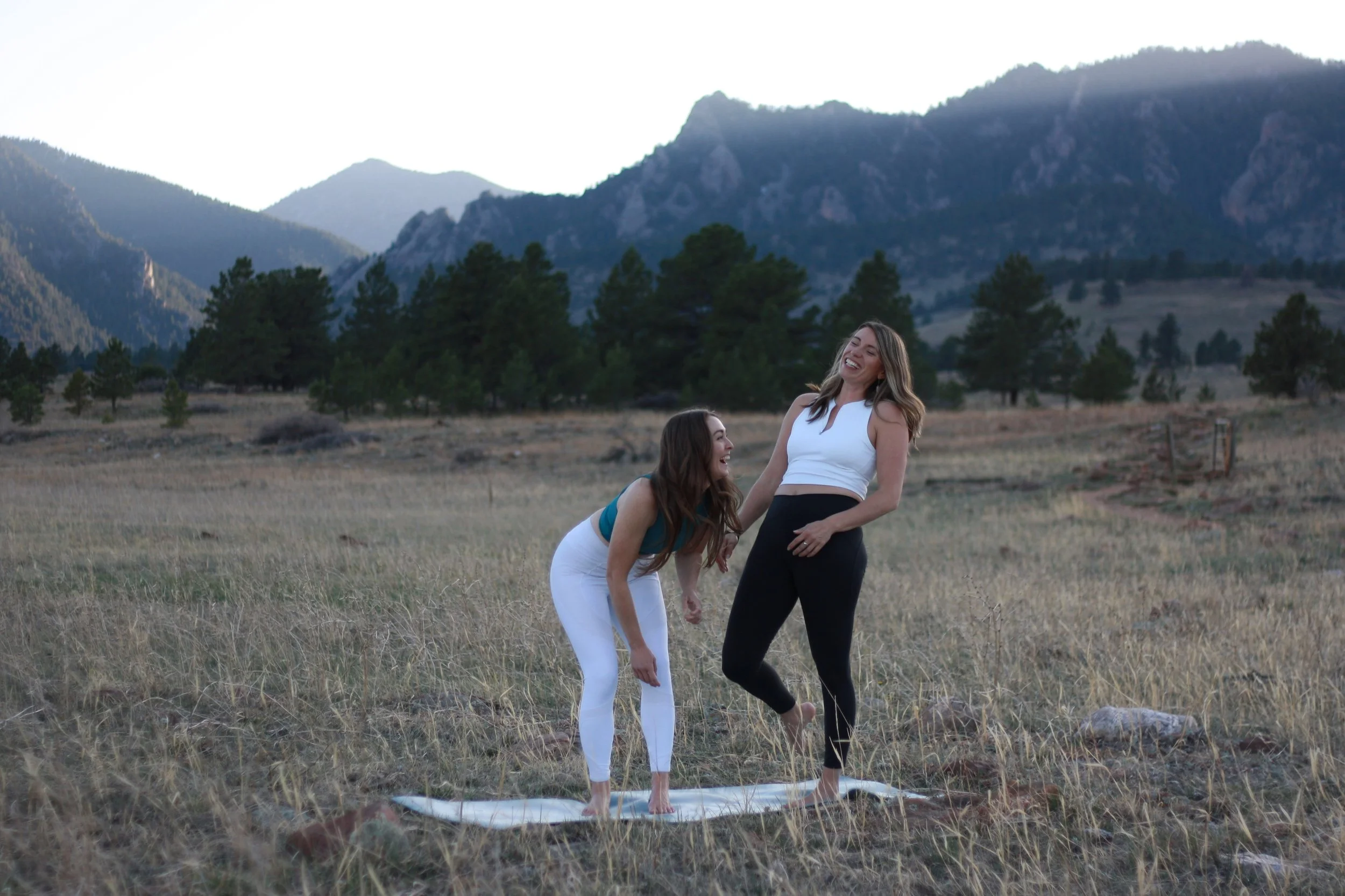 Two women practicing yoga on a mat in a grassy field with trees and mountains in the background, smiling and laughing.