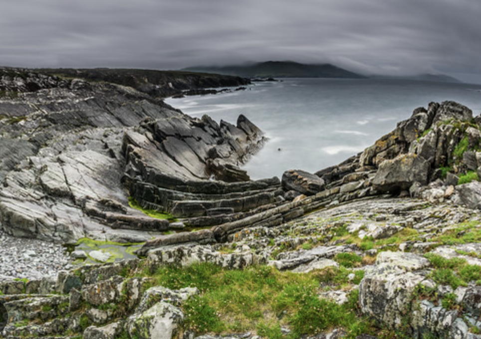 A rugged coastline with large grey rocks and boulders, patches of green grass and moss, and an ocean with waves under a cloudy sky in south Ireland.