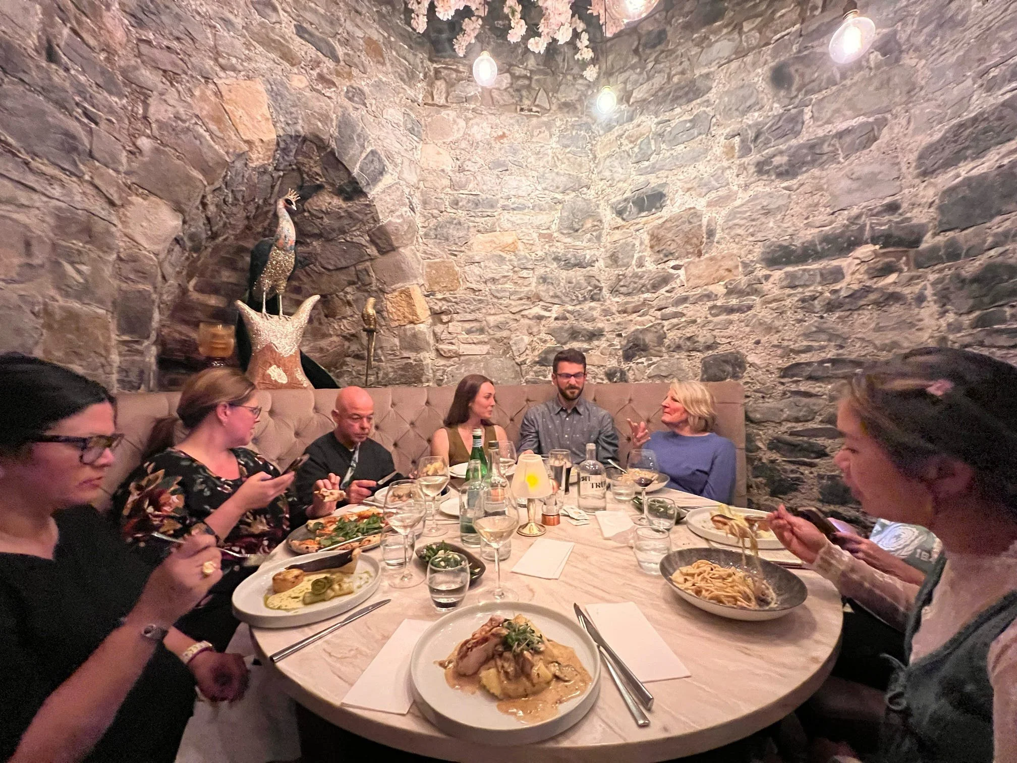 A group of people sitting around a round dining table in a stone-walled castle restaurant in Ireland, enjoying a meal at a yoga and art retreat. The table has food, drinks, and utensils, with some individuals engaged on their phones or talking.