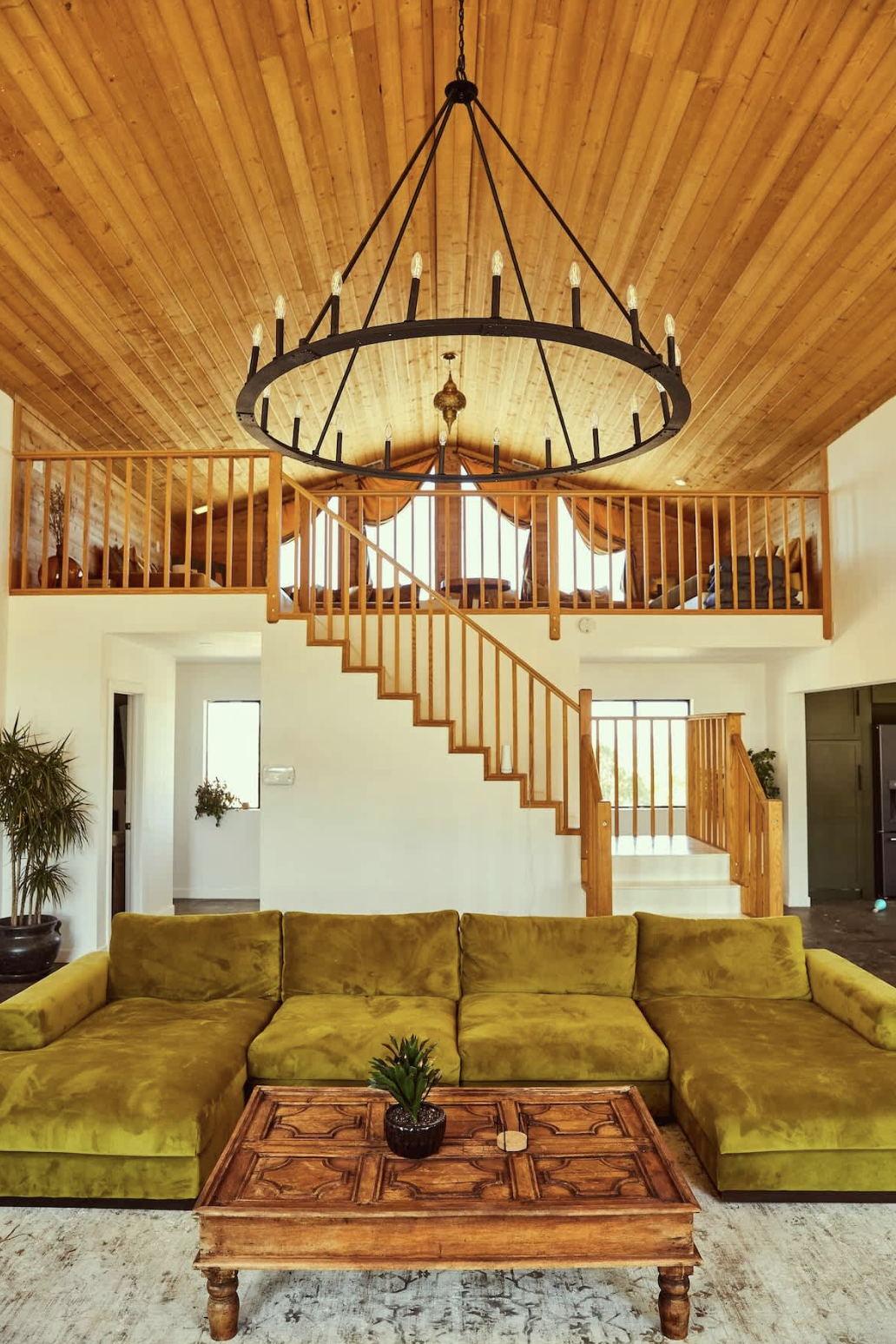 Living room with yellow sectional sofa, wooden coffee table with potted plant, staircase leading to a loft area with seating, wood-paneled ceiling, large circular chandelier with candle-style lights.