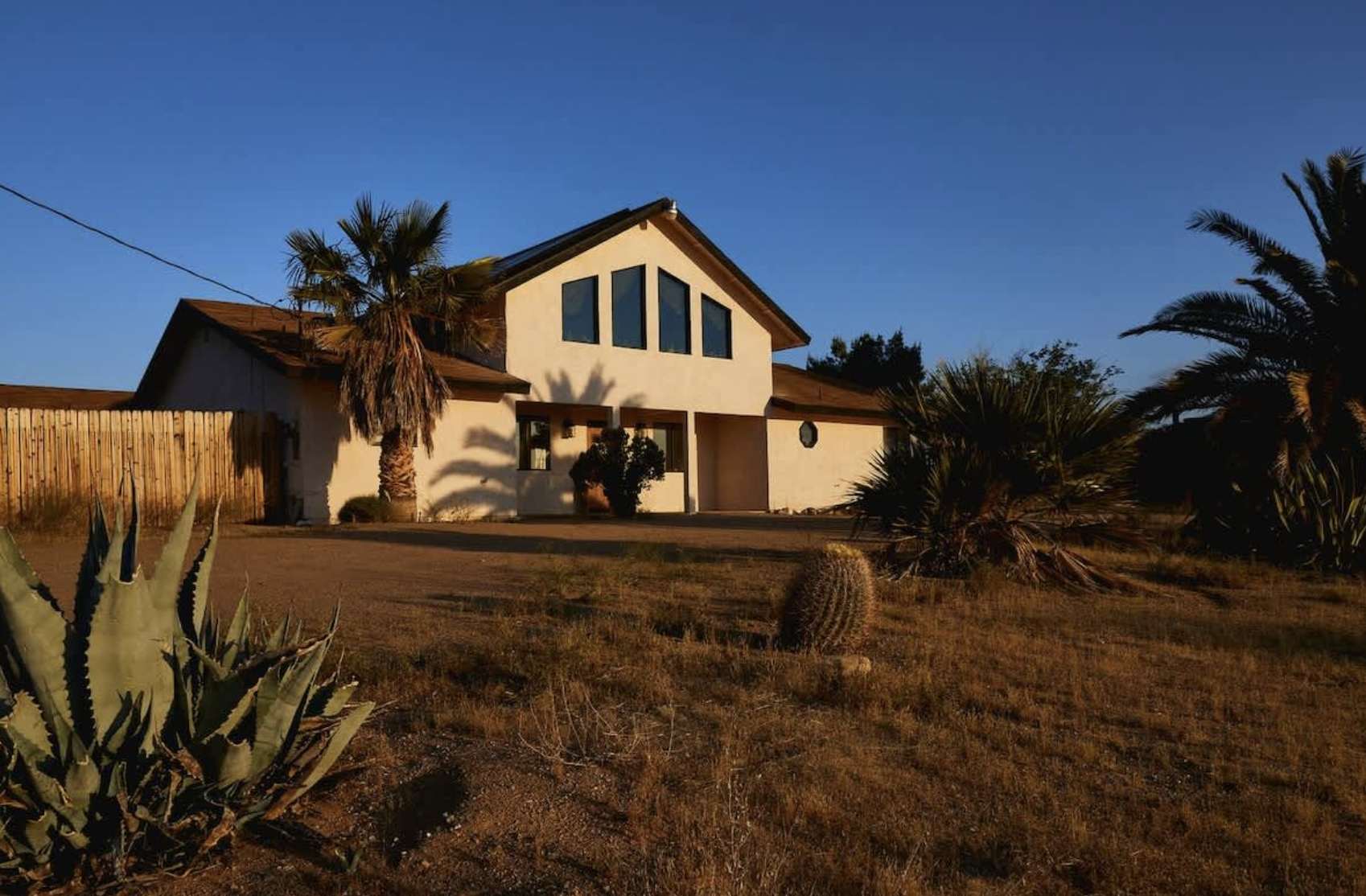 A white house with sharp, triangular windows, surrounded by desert plants and palm trees, under a clear blue sky during sunset.